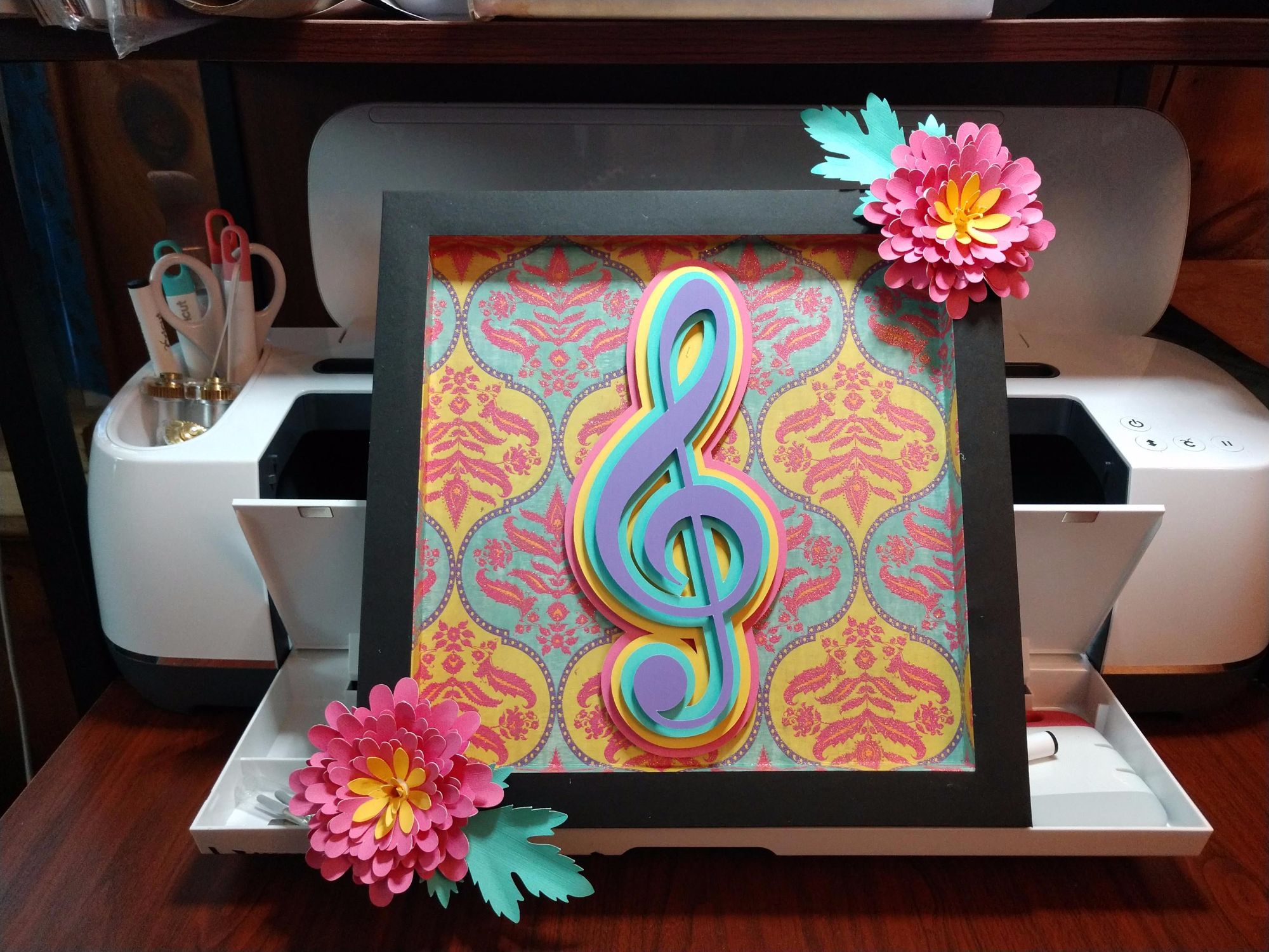Treble clef within a frame in bright colors of purple, teal, gold, and pink, with two large pink flowers at upper right and lower left on frame